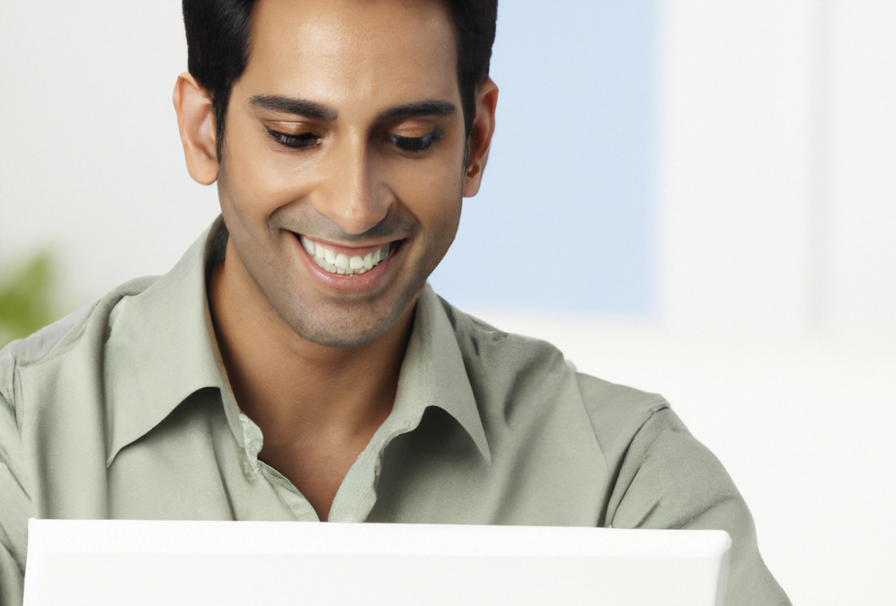 A South Asian person sitting in front of a laptop smiling.